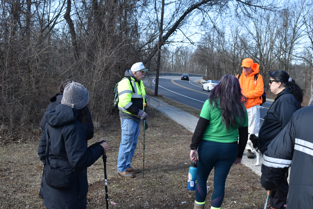 Eco tour guide speaking with participants along a roadside path near a wooded area.