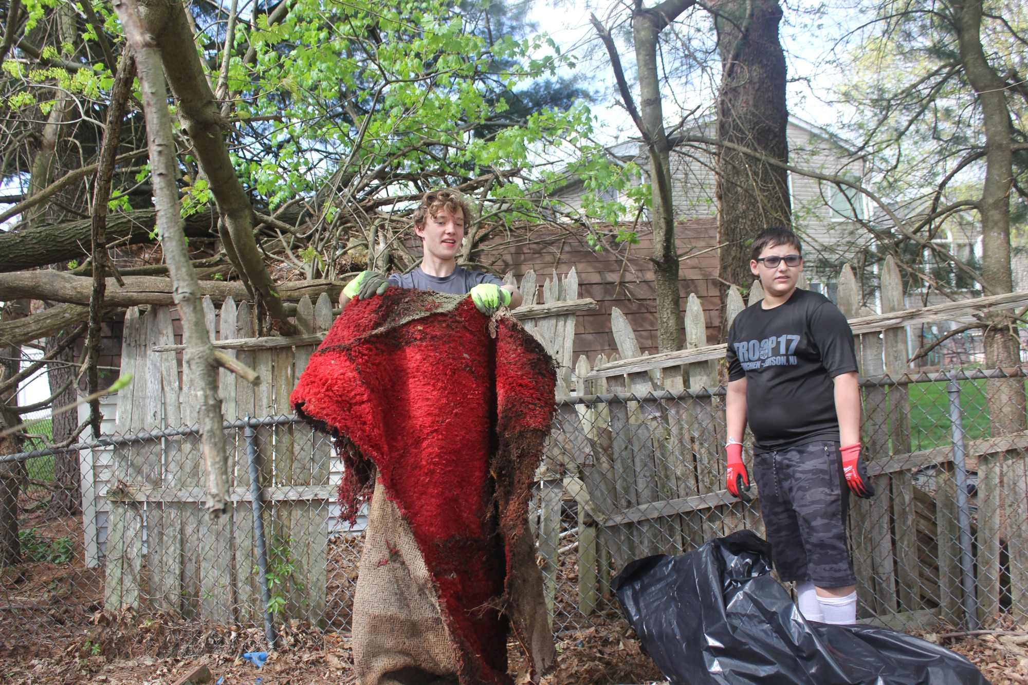 Scout holding up a dirty red rug beside another scout with a trash bag.