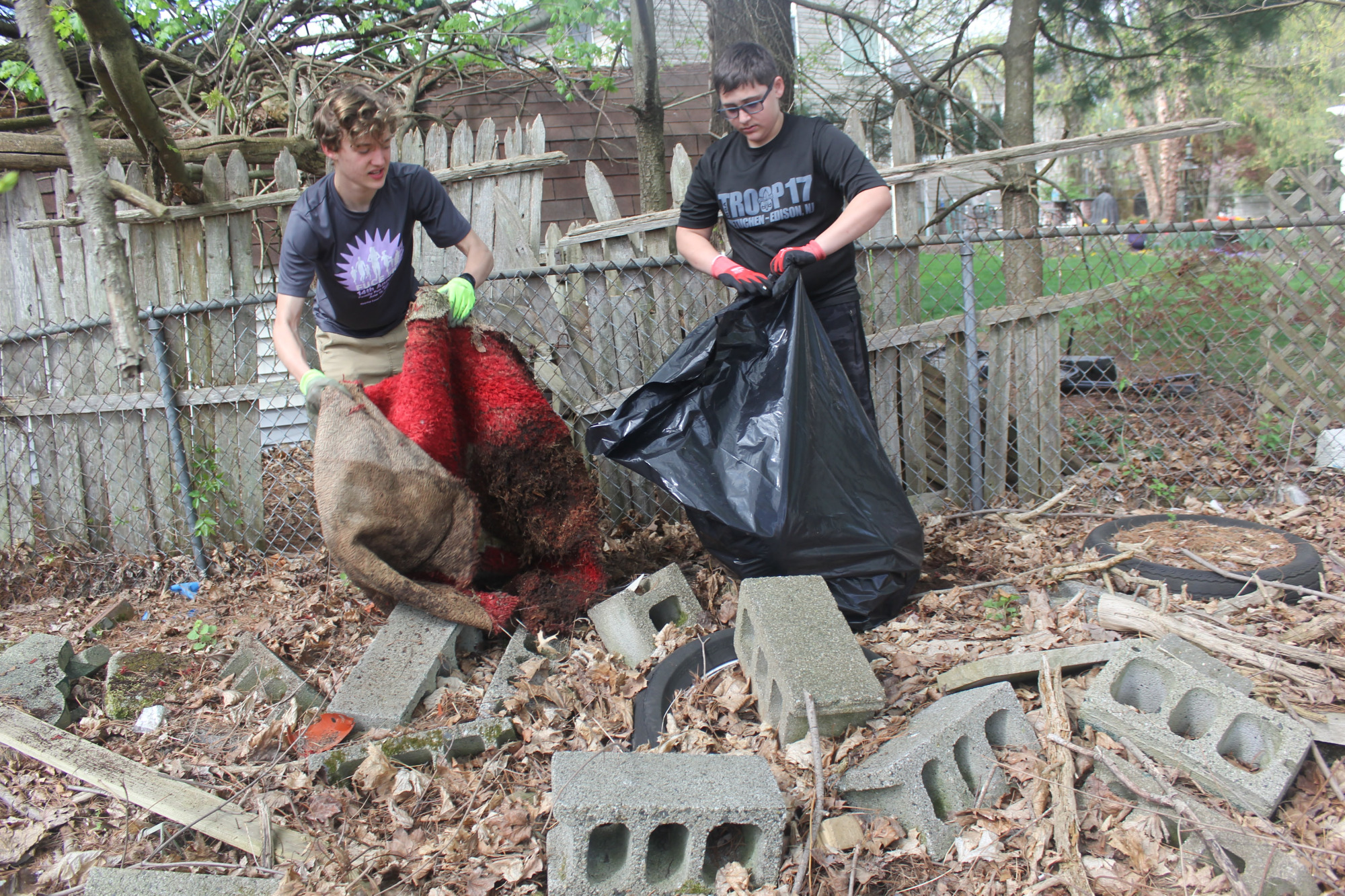 Scouts lifting a red carpet and placing debris into a trash bag near cinder blocks.
