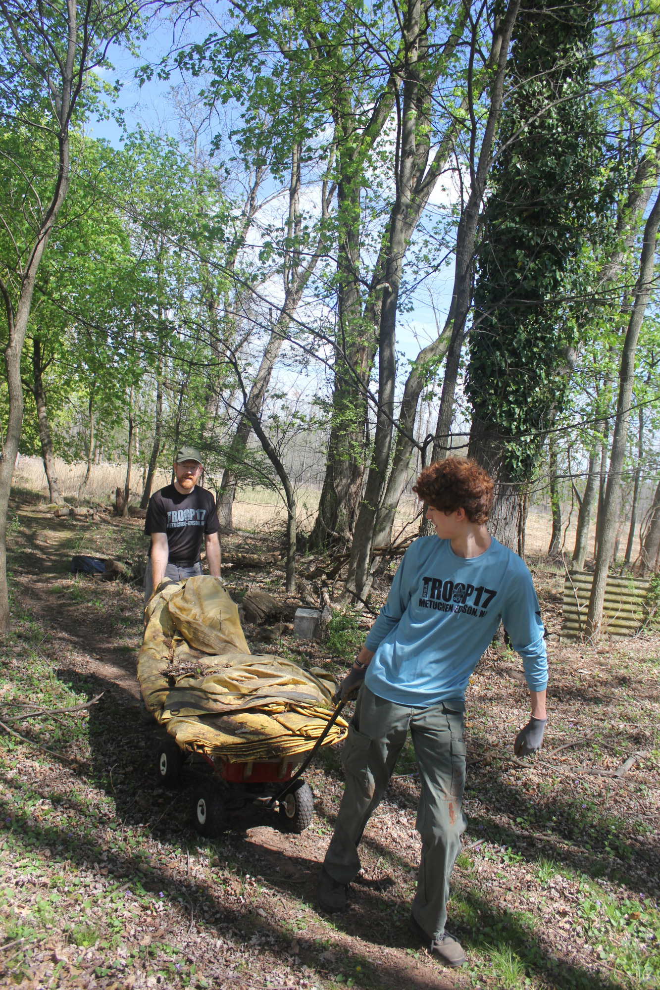 Scout pulling a cart loaded with bags of debris through a wooded area.