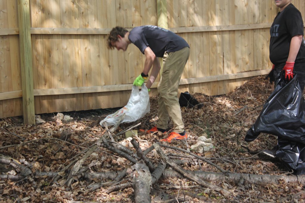Volunteer dragging a trash bag and bundled sticks near a fence.