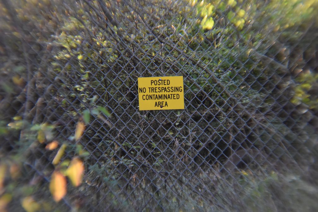 Yellow warning sign on a fence reading “Posted – No Trespassing – Contaminated Area” surrounded by dense vegetation.